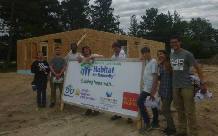 A group of people pose for a photo around a habitat for humanity sign. Behind them is the wooden frame of a house. 